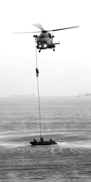 Black and white image of a sea rescue operation with a helicopter and lifeboat in Mumbai, India.