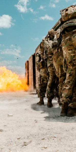 Group of soldiers in military gear facing explosion during training exercise outdoors.