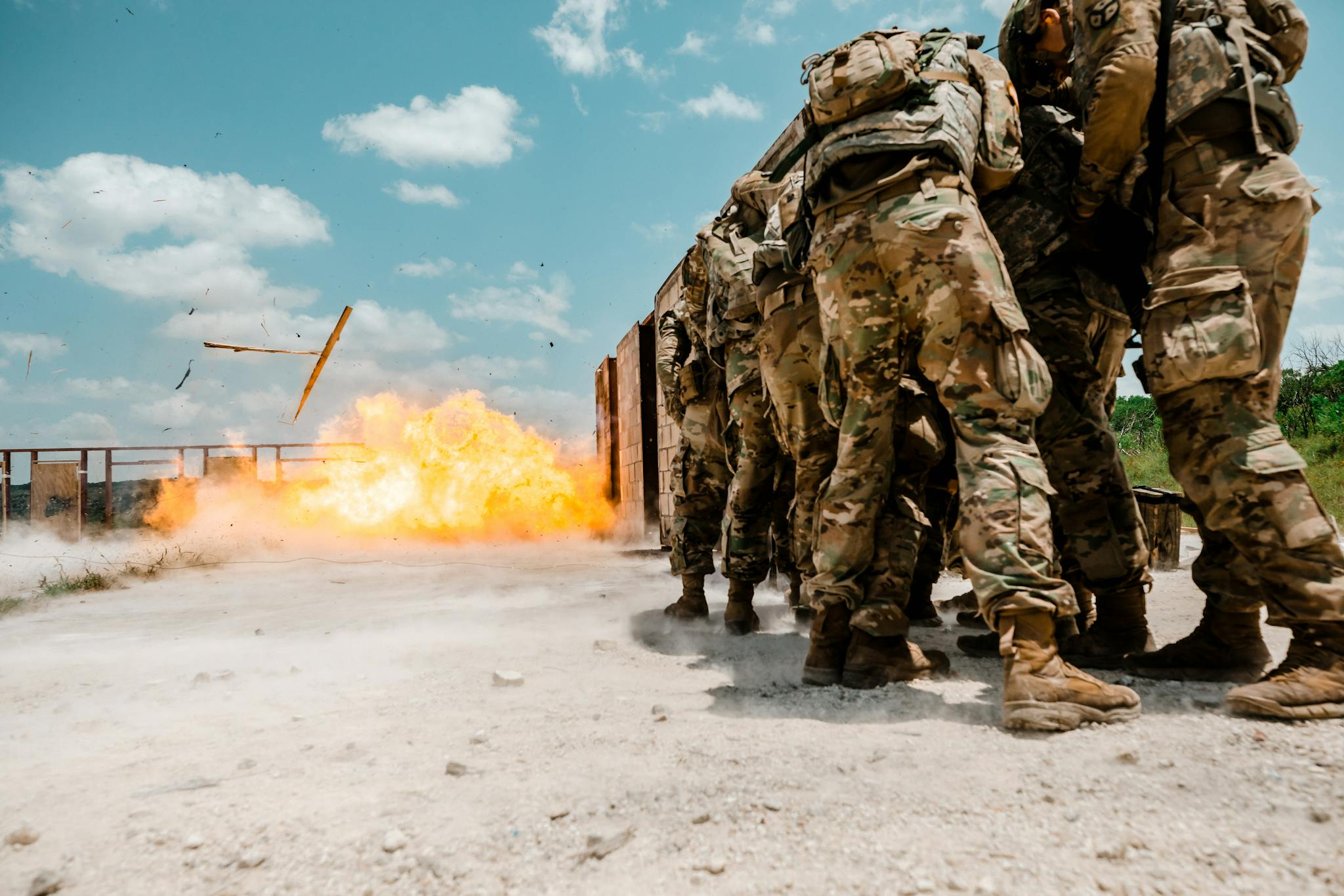 Group of soldiers in military gear facing explosion during training exercise outdoors.