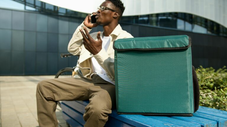 A delivery man sitting on a bench making a phone call with a package beside him.